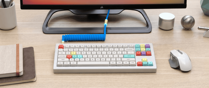 A modern white mechanical keyboard with colorful keycaps, connected by a coiled blue cable to a monitor on a clean wooden desk.