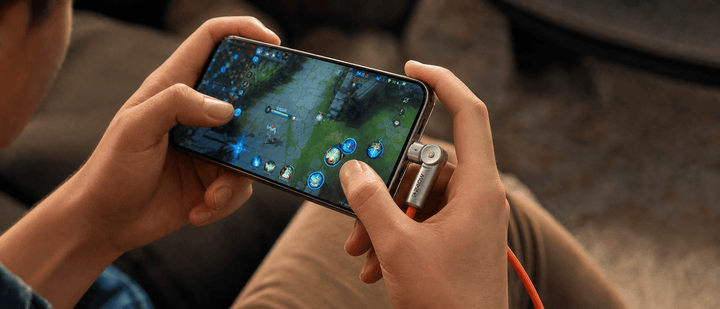 A person playing a mobile game in landscape mode while the phone is connected to an orange charging cable via a rotating USB-C connector.